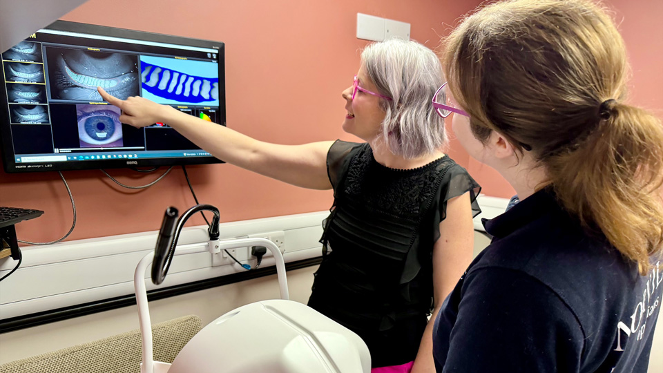 In a clinical room, Becci points at a screen displaying a meibomian gland assessment for dry eye, training a colleague who looks on with interest