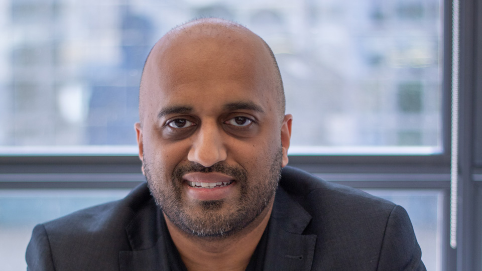 Mehul smiles with his hands folded on a table