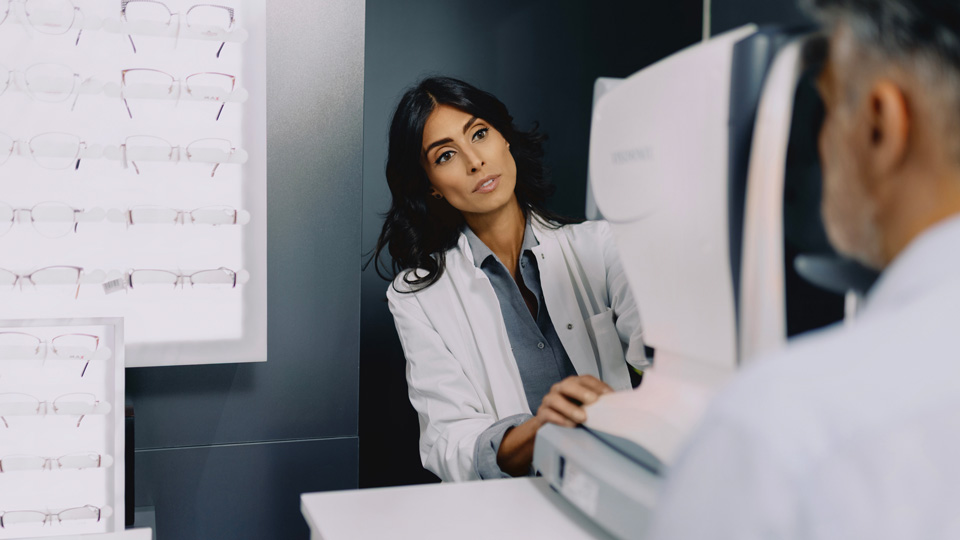 An optometrist carrying out an eye test on a patient. The optometrist is talking to the patient as leans around the device to do so
