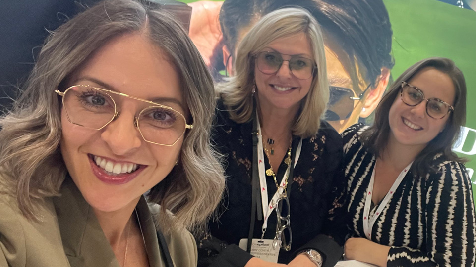 Three women gather around a table at an exhibition. Hayley, front left, wears a green suit jacket and gold wire RayBan style glasses. In the middle is Debby Coath in a black lace shirt and wire glasses