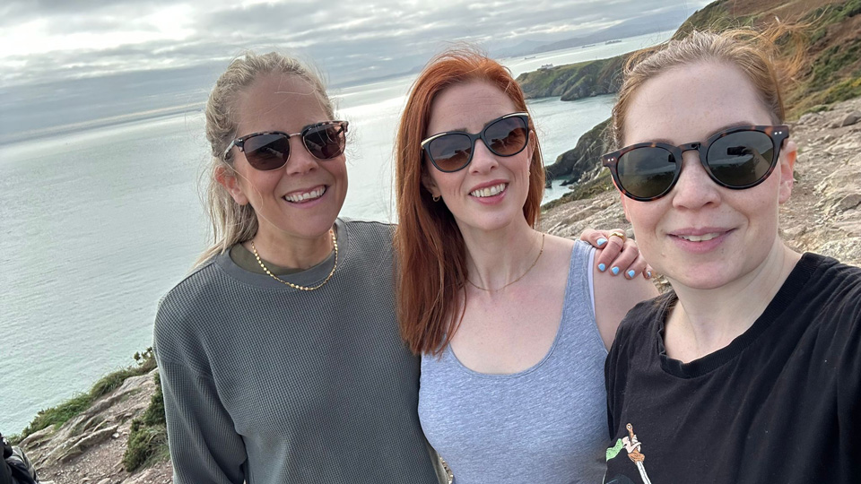 Three women stand on a rocky coastal path overlooking an ocean. The women pose together, all smiling and wearing sunglasses