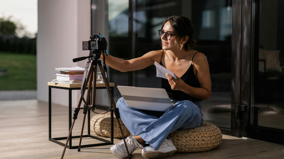 A woman sits cross legged on a sunny deck, setting up a camera on a tripod with a laptop balanced on her lap and notebooks around her