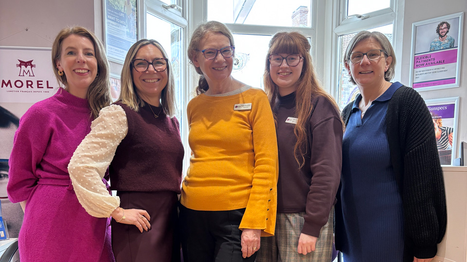 Five women stand together in the bay window of an optometry practice. Obe wears a bright pink woolen dress, beside her is a colleague in brown silk skirt, brown sweater vest as a long-sleeve blouse, in the center is Fiona Bromfield with a yellow bell-sleeve top, on the right is a woman in a long checkered light brown skirt, and on the far right is a woman in a navy dress with polo collar