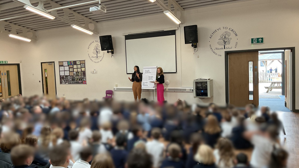 Two optometrists present with a paper flipchart to a school hall full of children, who are blurred out for privacy