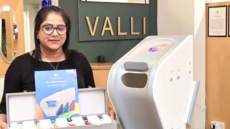 Nazmeen stands at the front desk to her Valli Opticians Blackburn practice, which is painted a deep grey-green with logos in brass and mirrors. On the desk beside her the colorimeter device has been presented. Nazmeen holds a boz in her arm of the different tints available