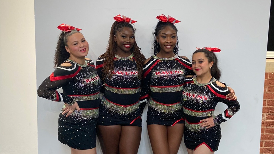 Four smiling young women stand together in a line, with arms around each other. They all wear cheerleading uniforms in black and red with silver diamante. They wear bright red silk bows in their hair.