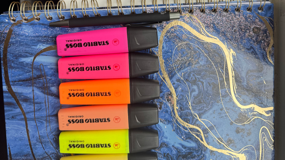 Arranged beside each other are an iPad, a dark notebook and pen, and a selection of bright highlighters