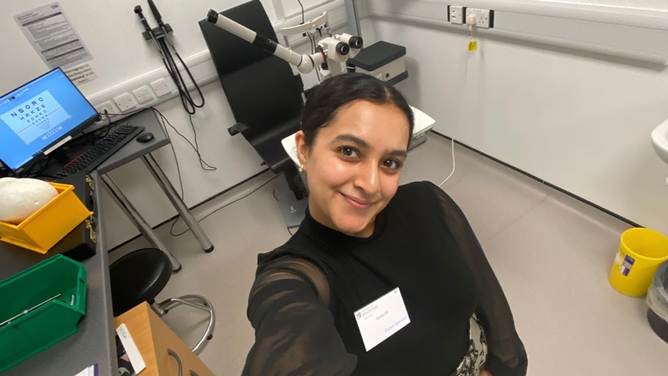 Komal takes a selfie from a high angle of the clinic room in practice. She wears a long sleeve black blouse with a patterned white skirt. Behind her is the Snellen chart and testing equipment
