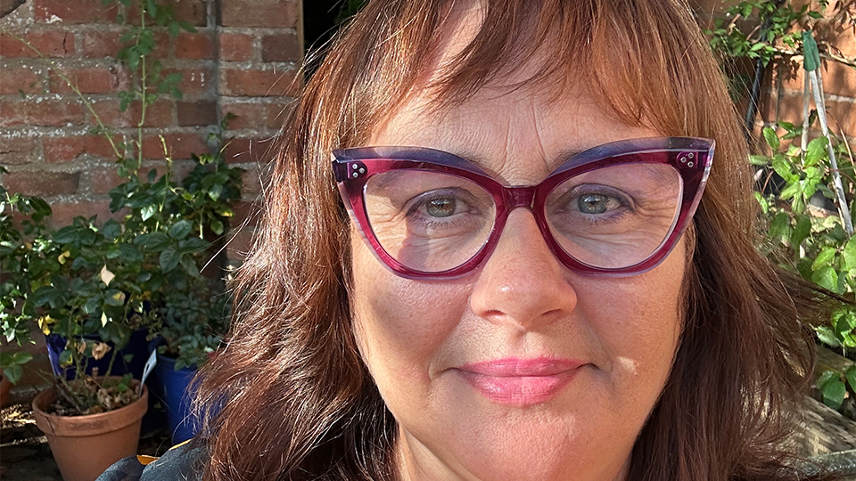 Portrait of a woman wearing burgundy cat-eye glasses and a colourful patterned blouse, photographed outdoors in front of an ivy-covered brick wall