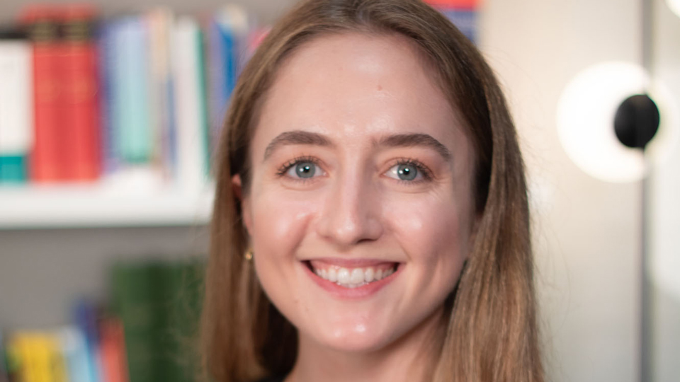 A smiling woman with long brown hair wearing a black dress and necklace, standing in front of a blurred bookshelf