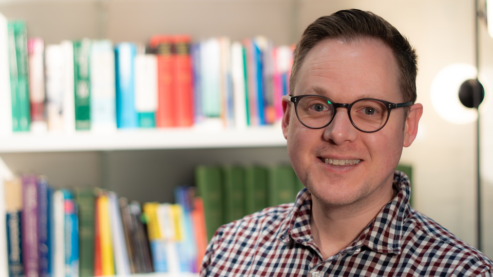 A smiling man wearing glasses and a checked shirt, standing in front of a blurred bookshelf