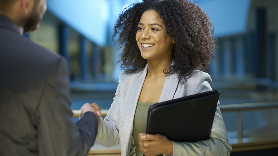 A professional woman shakes hands with an interviewer while holding a folder