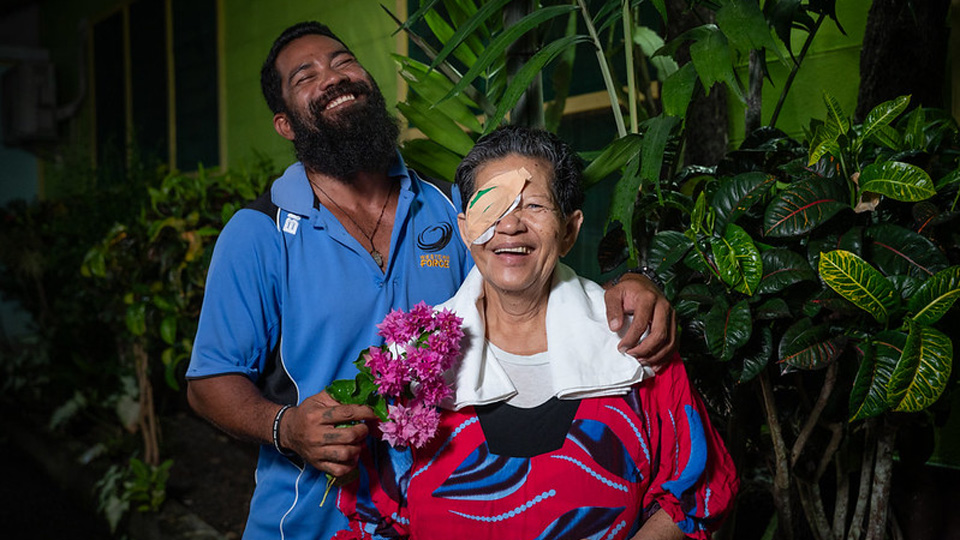 An older woman and younger man are laughing happily together. The woman has a patch over one eye, suggesting she has had cataract surgery, while the man holds her shoulders in a friendly manner