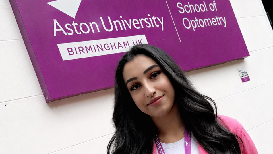 Umamah stands beside the purple sign for the Aston University School of Optometry