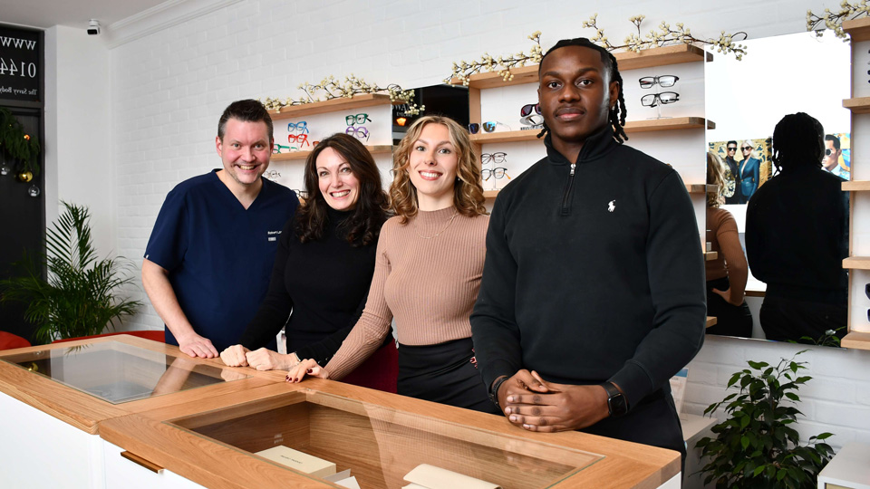 A team of four smile behind a display table in an optometry practice. The environment is warm and airy, with frames behind them and dried flowers for decoration