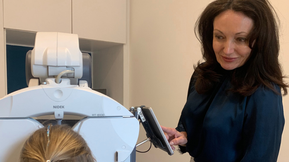 Claire Hayden performs a vision assessment on a child who has their back to the camera. Claire has shoulder-length curled brown hair and is dressed in a navy silk blouse and white trousers