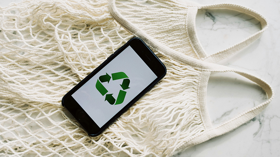On a marble worktop a crocheted shopping bag is spread out. A smartphone is laid on top with the universal logo for recycling, three green arrows in a triangle loop, displayed