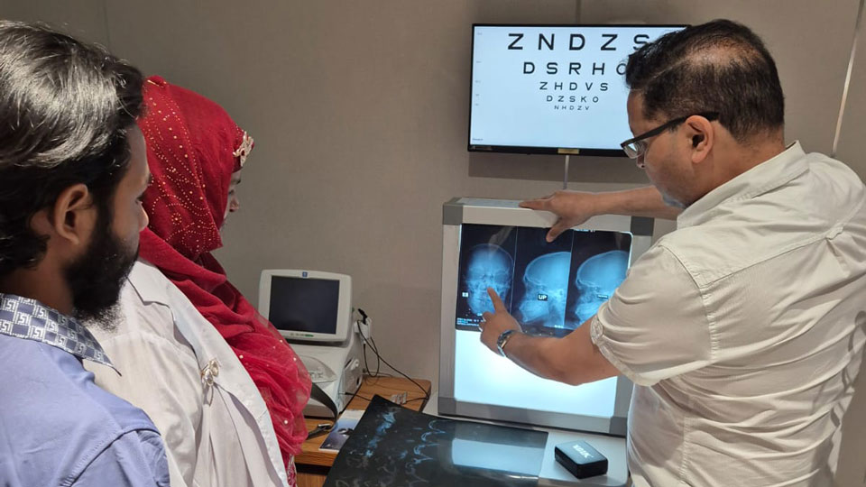Niaz is pointing to x-rays on a display board, underneath a snellen chart. Two people, one in a white coat, look on.