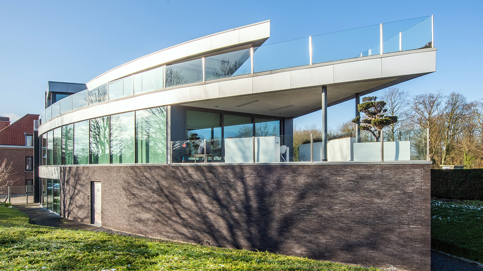 A modernist building made of grey brick, glass and metal, reflecting the bright sunshine. Snow on the ground and the shadow of a bare tree indicate a cold climate or winter season, though a bright blue sky caps the scene