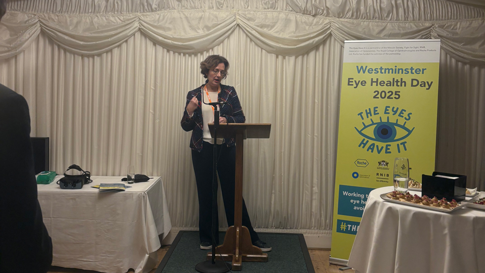 Alt text: A woman stands at a lectern speaking into a microphone at an indoor event with white draped walls and a chandelier overhead; a green banner beside her reads “Westminster Eye Health Day 2025” with an eye graphic, and tables with information materials and refreshments are positioned around the room.