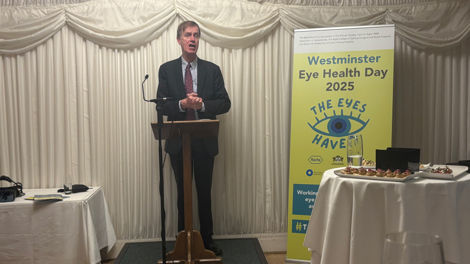 A man in a dark suit stands at a lectern speaking into a microphone in a small event space with white draped walls; beside him is a green banner reading “Westminster Eye Health Day 2025” with an eye graphic, and tables with refreshments are visible