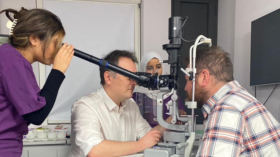 A hospital optometrist is using a slit lamp to perform a sight test on a patient. A student in purple scrubs stands to the side and looks down a viewpiece to see what the optometrist is seeing. Another student observes across the room