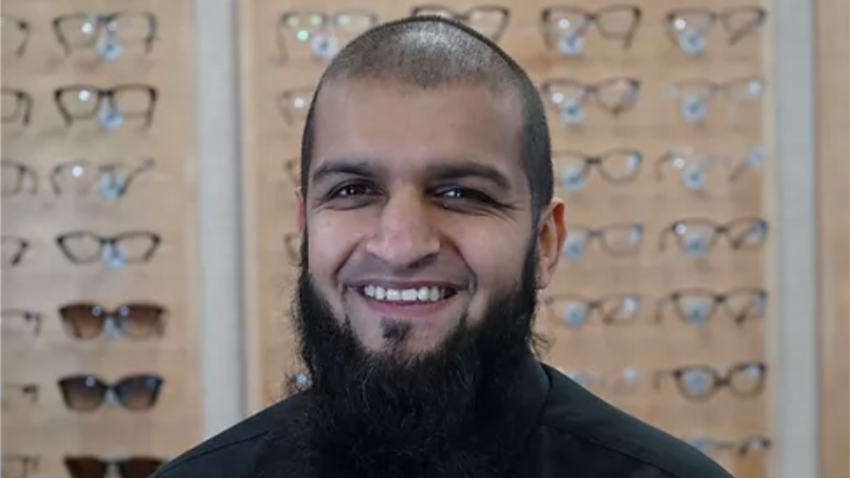 Sohail smiles wearing a black shirt and tie in front of a backdrop of glasses
