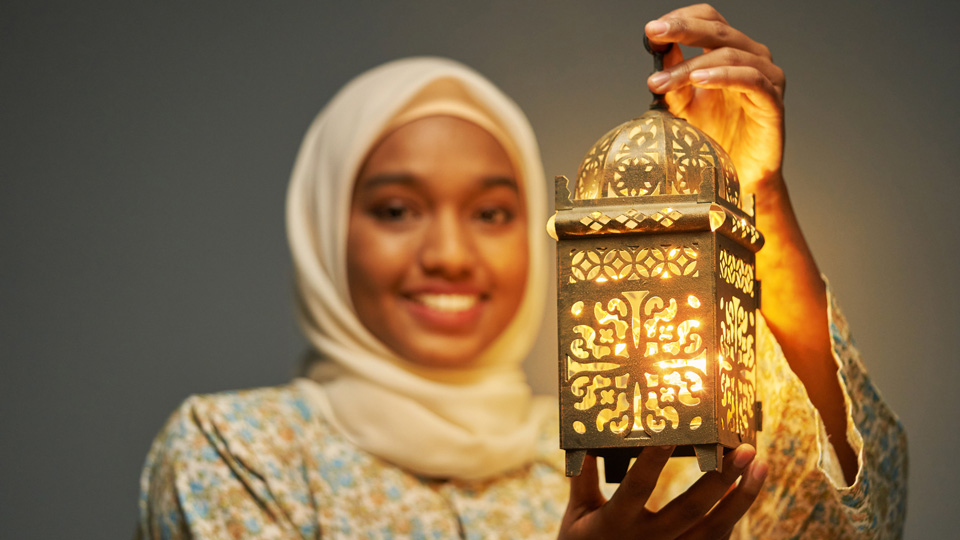 A woman in a white hijab holds a gold lantern in front of her