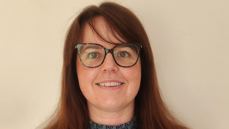 Rachel wears a blue and black patterned blouse and has large dark tortoiseshell spectacles