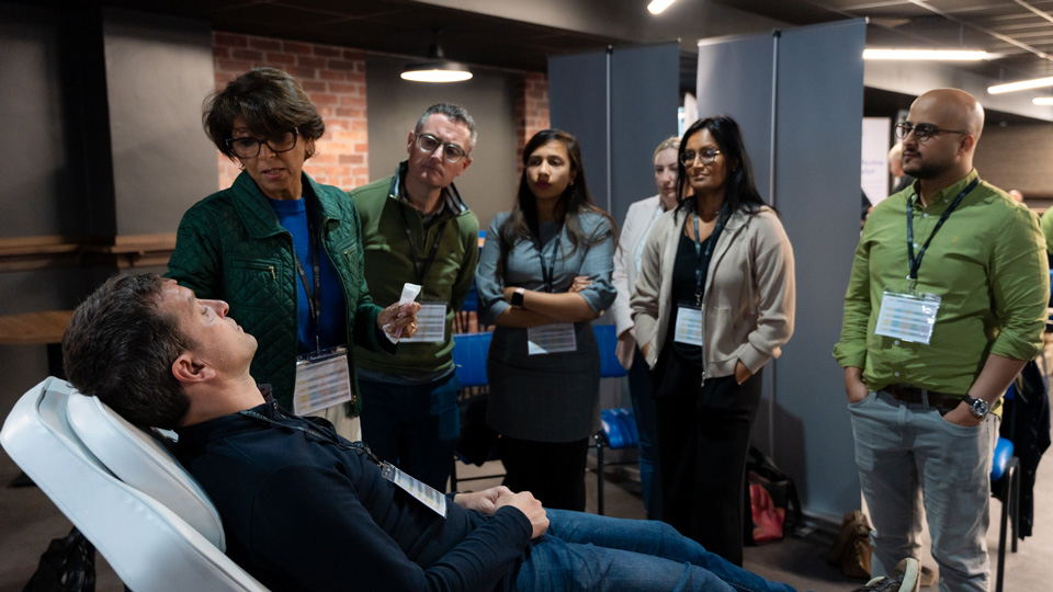 A crowd watching a practical dry eye demonstration, stood next to a man laying down on a white chair