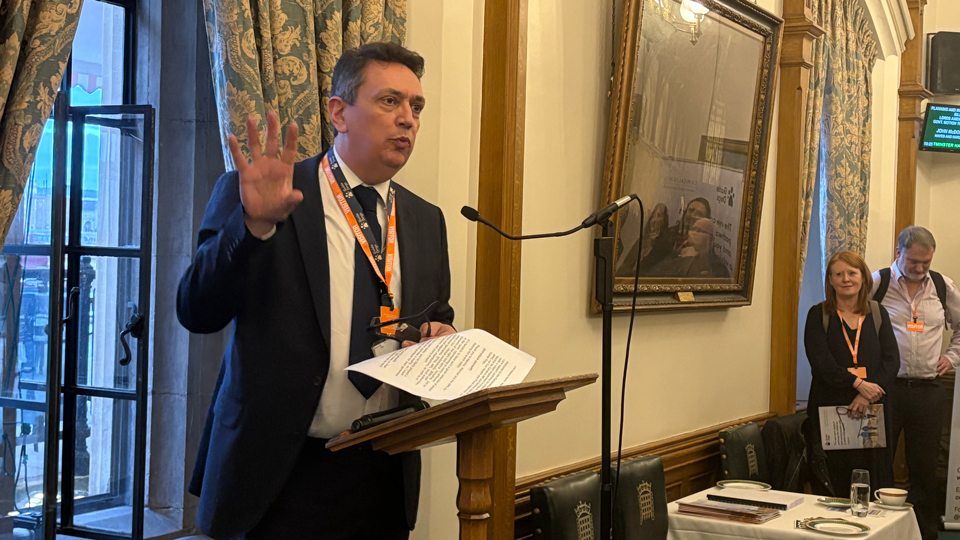 Andrew Lennox, the CEO of Guide Dogs, is wearing a suit and stands at a lectern giving a speech in an ornate room with tall, patterned curtains and wood panelling. He gestures with one hand while holding papers in the other. A microphone is positioned in front of him, and a few people stand in the background listening. Chairs with a crest emblem and a table set with plates and glasses are visible nearby.