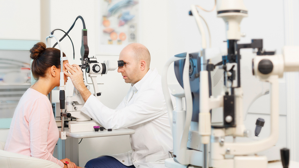 In the foreground is a piece of optical equipment. In the background, an optometrist checks the eyes of a patient
