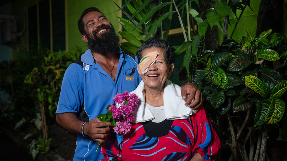 Makalika and her son are posed together, laughing. Makalika wears a colourful red and blue patterned top and has an eye patch following surgery. Her son holds a handful of bright pink flowers, and his head is thrown back in a laugh 