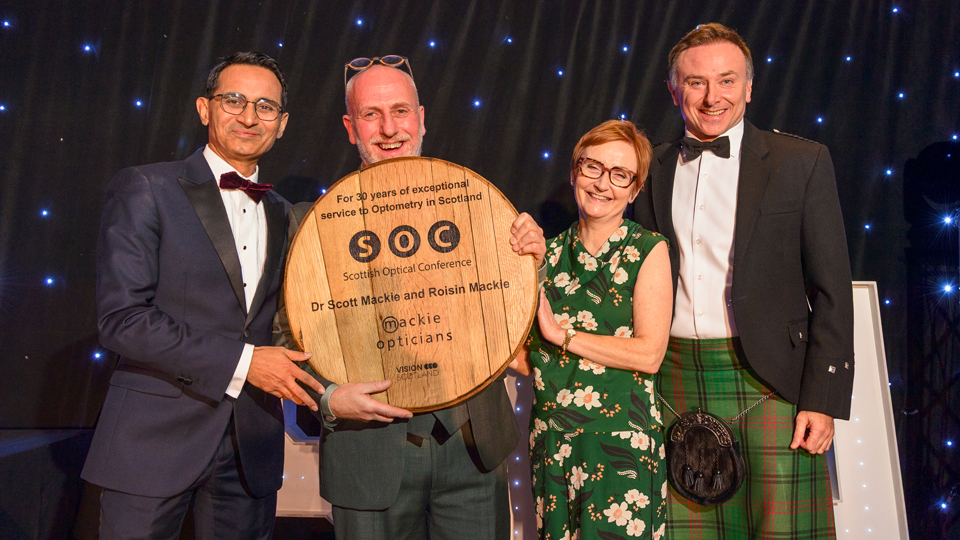 Dr Scott and Dr Roisin Mackie smile around a wooden plaque the size of a serving plate that celebrates 30 years of providing CPD. The pair look surprised but thrilled. Two people join them on stage to present the award