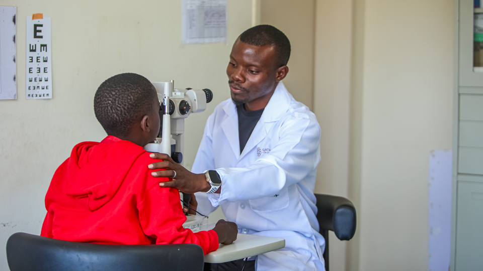  A healthcare professional in a white lab coat conducts an eye examination for a child wearing a red hoodie using a slit-lamp machine in a clinic setting. An eye chart hangs on the wall in the background