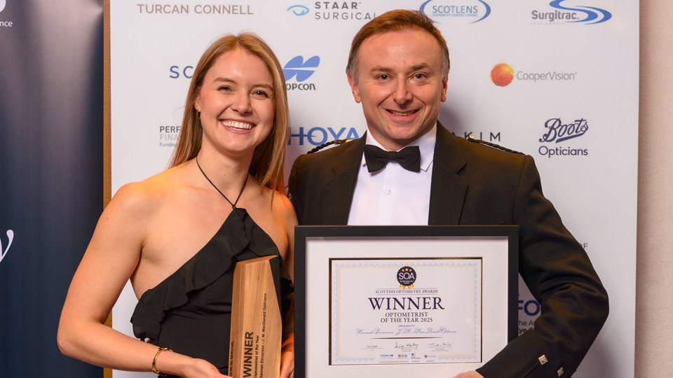 Hannah Einarson smiles broadly at the camera, she wears a black halterneck dress and holds a wooden award. Beside her, Jonathan Ross, from Vision Scotland, presents the certificate