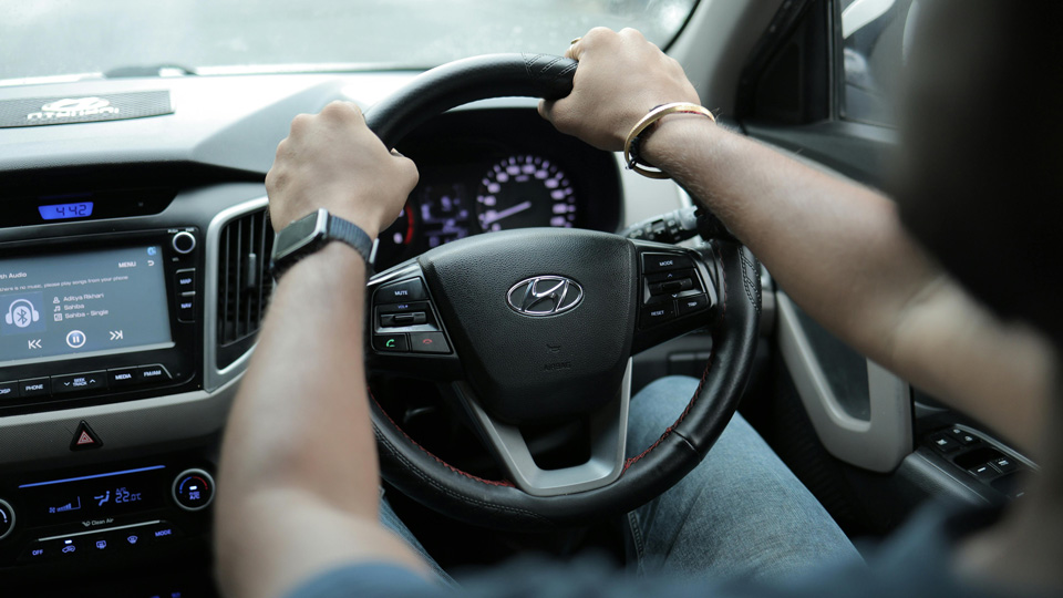 A driver’s hands on the wheel of the car in the 10 and 2 position. The photograph is taken over the driver’s shoulder and focused on their hands