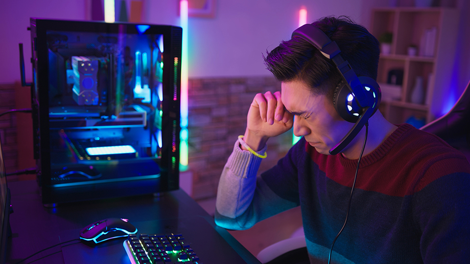 A man sits in a darkened room next to a computer screen rubbing his eye