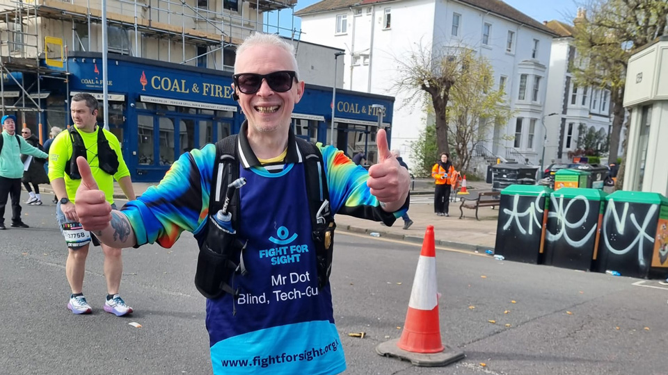 Clarke, wearing blue and yellow running attire and a Fight for Sight running bib, poses with thumbs up while running. He wears Meta smart glasses