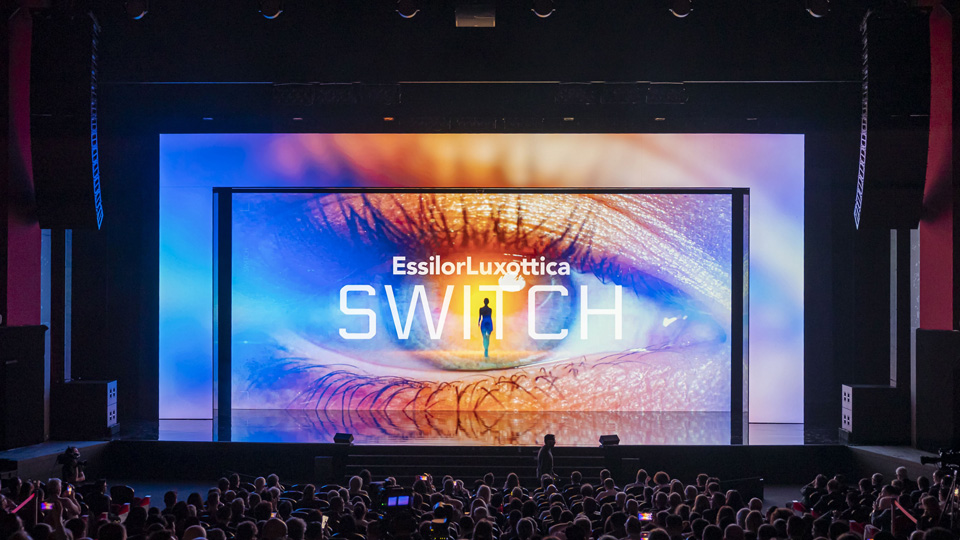 A huge screen behind a stage displays a close-up of an open eye, with a wash of blue light on either edge. Text reads EssilorLuxottica Switch, and in the centre of ‘Switch’ – also the centre of the photo of the eye – is a robot-like figure. The overall impression is one of futuristic eye health