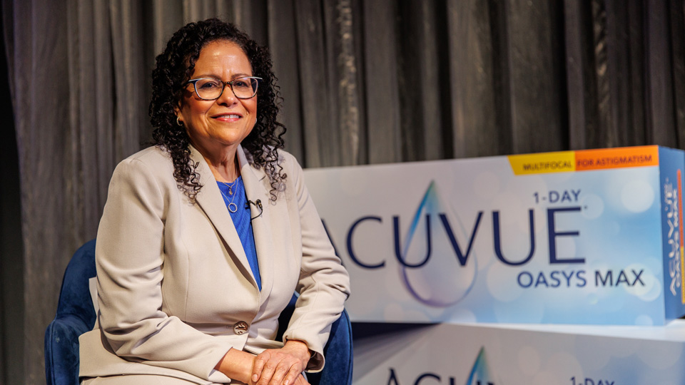 Dr Giovanna Olivares is seated beside two giant boxes designed to resemble a contact lens box. She wears a tan-grey suit with a bright blue top and cat’s eye glasses