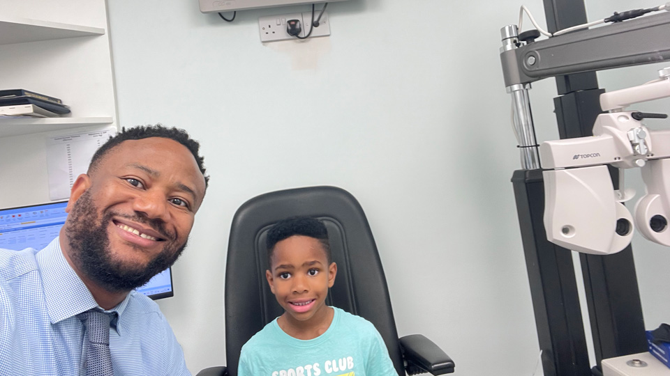 A smiling optometrist takes a selfie with a young child seated in an eye examination chair. Behind them, a digital eye chart is mounted on the wall, and ophthalmic testing equipment is visible to the side in a clinical room