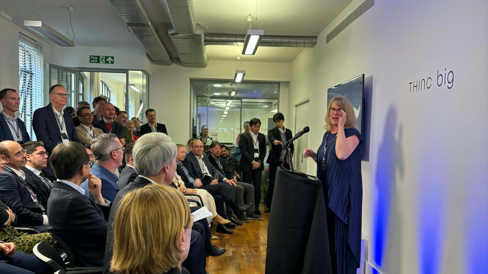 Krysten Williams, wearing a royal blue pleated two piece set, stands at a lectern to welcome attendees to the THINC grand opening