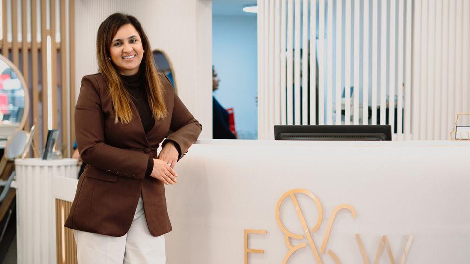Dr Pretty Basra leans an elbow against the white and gold reception desk of her practise, which displays the cursive E&W Opticians logo. Pretty has brown hair past her shoulders, a deep brown suit jacket and white trousers