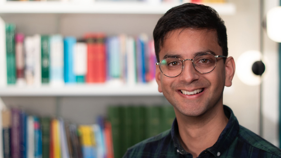 Karan is photographed in an office library. He wears a navy and deep green plaid shirt he has thin-rimmed metal glasses