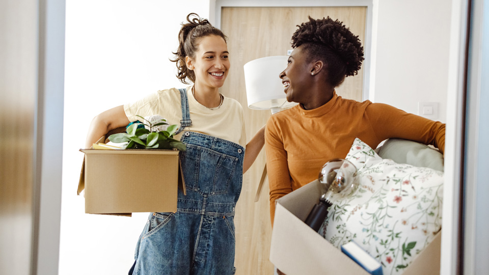 Two students carry cardboard boxes filled with small items and furnishings into an apartment, they are laughing and happy