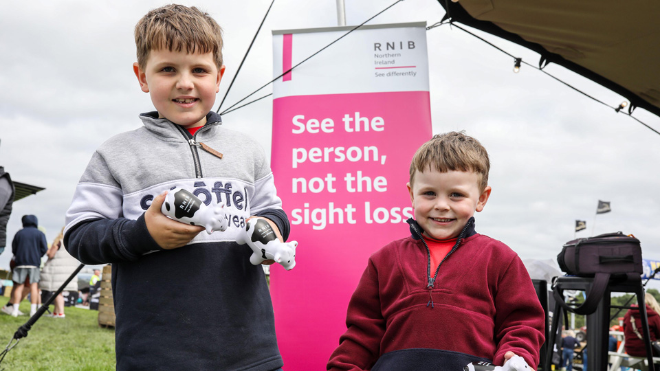 Two, small, smiling boys holding RNIB cows beside the RNIB pop up