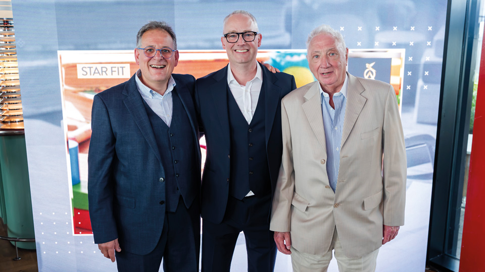 Three men in business attire stand in front of a light screen, celebrating eighty years of Millmead Optical Group