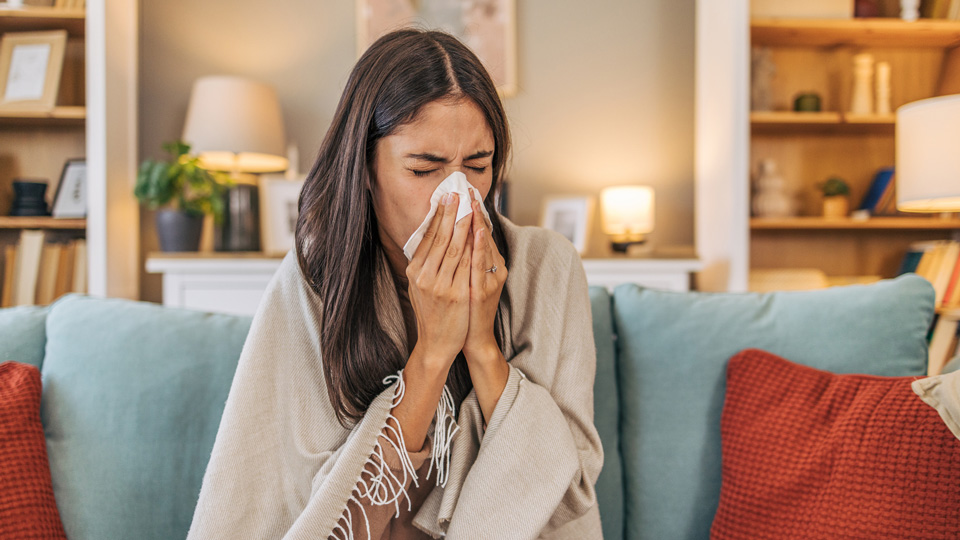 A woman sits on the sofa wearing a blanket and blowing her nose