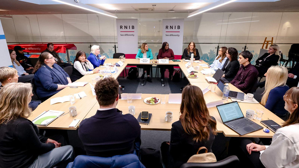 A wide shot of a roundtable meeting with around 20 people seated at U-shaped tables in a modern conference room. Two RNIB banners reading “See differently” stand behind the speakers at the head of the table. Attendees listen and take notes, with laptops, notepads, mugs, and plates of fruit on the tables